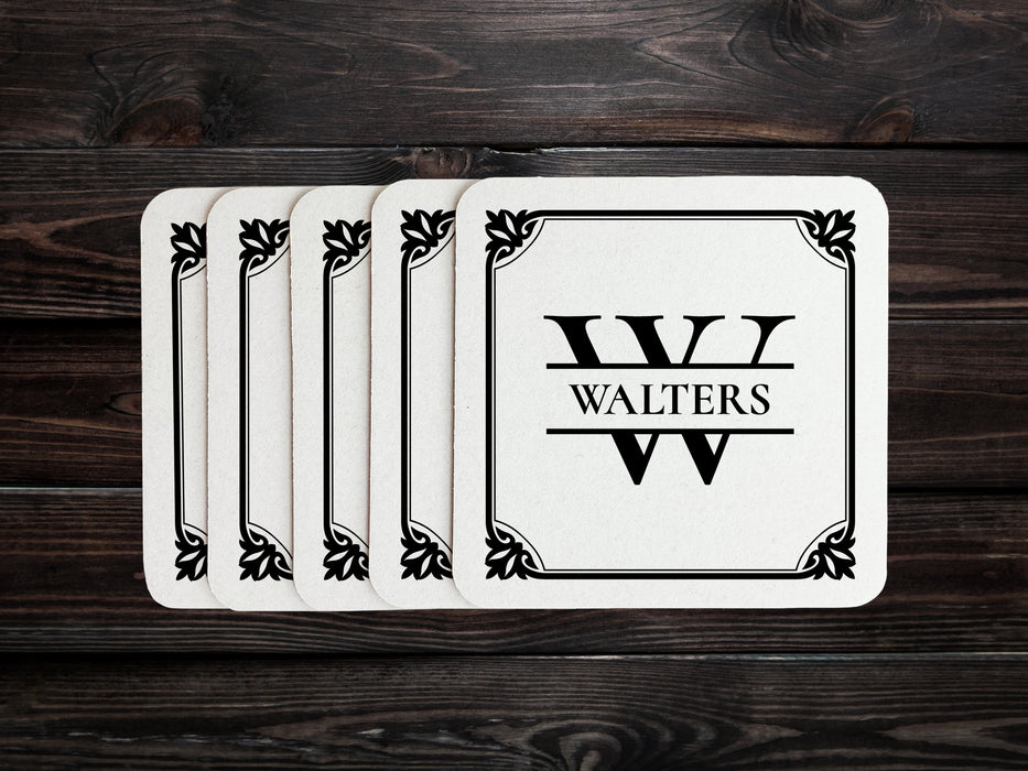 A stack of coasters are spread out on a dark wooden table. Coasters feature Framed Monogram Family Name design. Design has a square ornamental frame around a monogram for the last name "Walters". Design is printed on a white coaster.