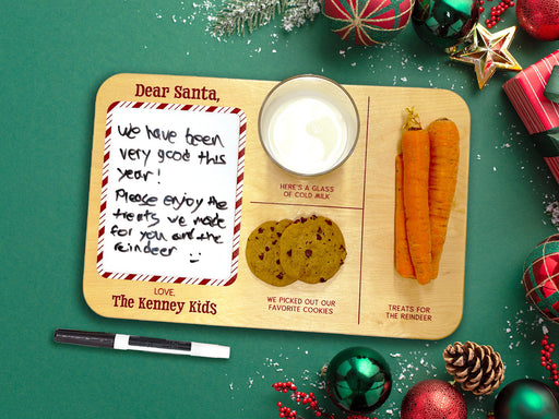 wooden Santa cookies and milk tray with a glass of milk, cookies, and carrots on top of a green background next to a dry erase marker surrounded by holiday decor such as pine leaves, holly berries, and christmas ornaments