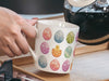 woman holding a 15 oz white ceramic mug with an easter pattern of floral decorated eggs with a baby chick popping out of an egg in a kitchen pouring a pot of coffee