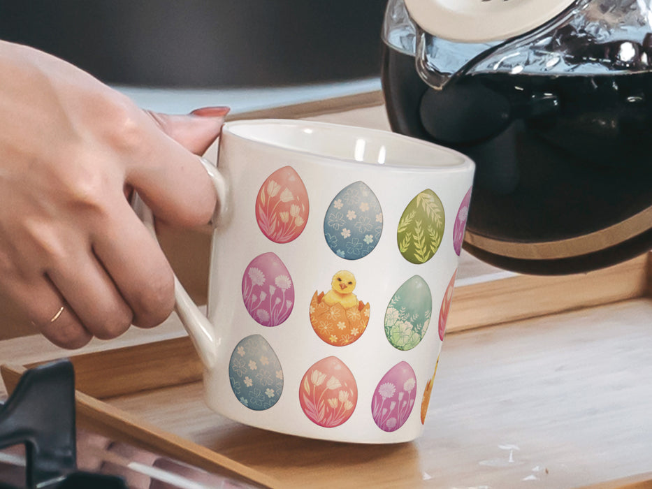 woman holding a 15 oz white ceramic mug with an easter pattern of floral decorated eggs with a baby chick popping out of an egg in a kitchen pouring a pot of coffee