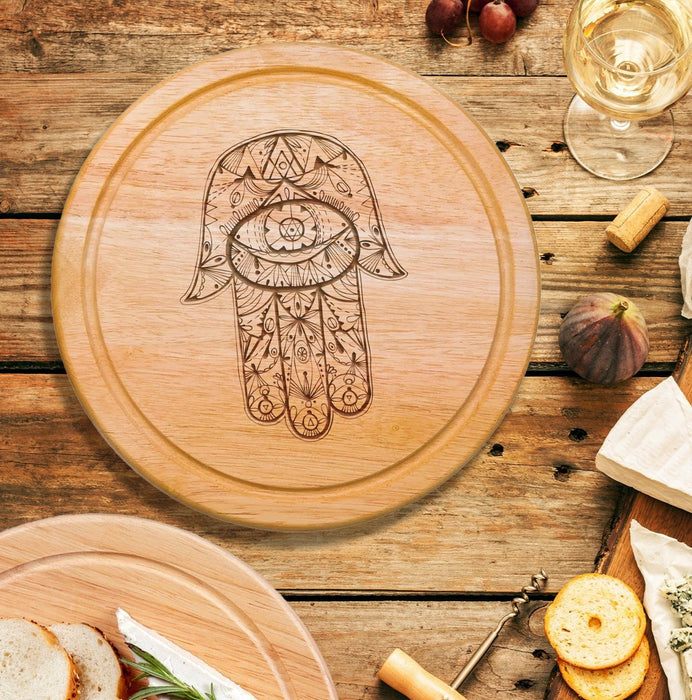 wooden cheeseboard on wooden table surrounded by wine glasses, figs, cheese, bread, crackers, Italian meats, and another charcuterie board engraving includes a Hamsa design