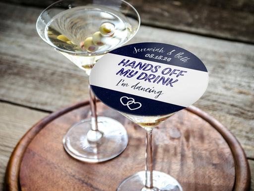 Don't Take My Drink coaster placed on top of martini glass on round wooden tray. Coasters say married couple names, wedding date, HANDS OFF MY DRINK I'm dancing. Coaster design is printed in purple and black on a white coaster.