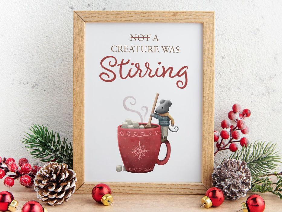 wooden frame with christmas print of mouse stirring hot chocolate with marshmallows and typography ontop of wooden countertop surrounded by winter decor such as pinecones, pine leaves, holly berries, and red ornaments