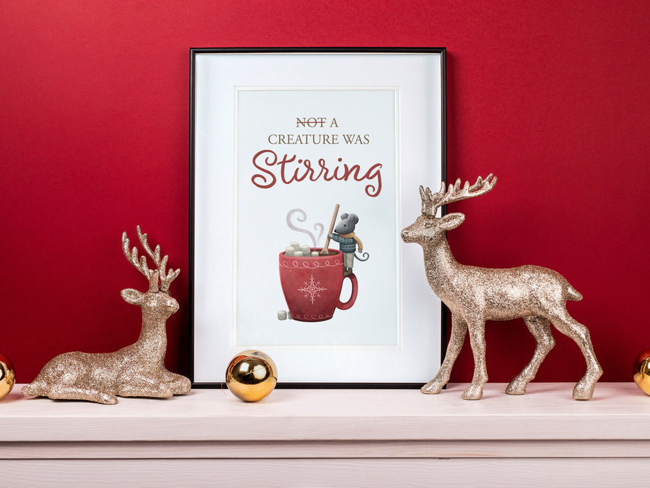 large frame with a christmas print of a mouse stirring hot chocolate with marshmallows and typography  against a red background ontop of a white wooden countertop surrounded by holiday decor such as golden ornaments and gold sparkly reindeer