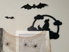 A black door frame topper, designed with the silhouette of a howling wolf, full moon, and clouds, is seen on top of a white picture frame. The frame is covered in spider webs and fake spiders. The beige wall behind has fake bats attached to it.