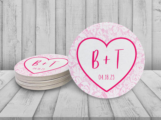 A stack of coasters by a single coaster on white wooden background. Coasters feature Heart and Lace design. These are designed with the bride and groom first name initials and wedding date surrounded by a hot pink heart outline and light pink lace.