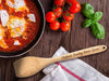 flat wooden spoon next to a pot full of sauce ontop of wooden table with engraving on the handle that says Chelseas Sunday Sauce Spoon next to tomatoes and basil