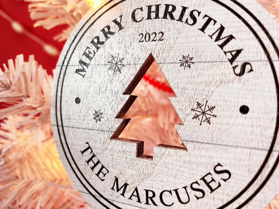  Ornament shown hanging from a Christmas tree. The ornament is wooden and shaped like a circle with a Christmas Tree cutout in the middle. Ornament says Merry Christmas, Year, and Family Name