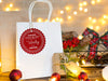 A red glitter cardstock Santa gift tag is shown hanging on a white gift bag. The bag is sitting on a wooden table, surrounded by lights, Christmas ornaments, and other Christmas elements.