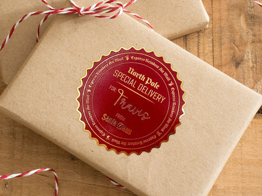 A gold foil cardstock Santa gift tag is shown on a craft paper wrapped box. The box also has a red and white striped string on it. The box is seen on a wooden surface.