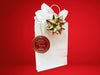 A gold foil cardstock Santa gift tag is shown hanging on a white gift bag. The bag is on a red backdrop and has a gold bow on it.