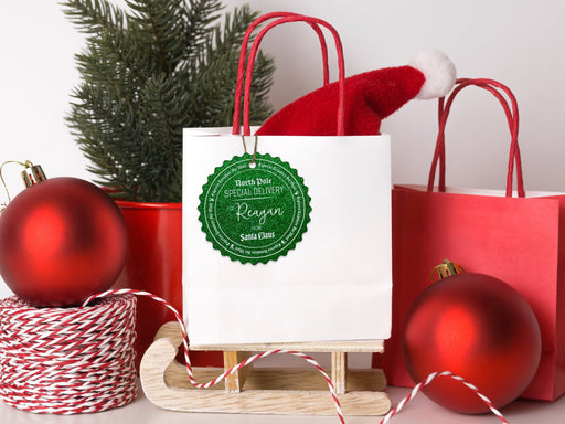 A green glitter cardstock Santa gift tag is shown hanging on a white gift bag. The bag is sitting on a sled and is surrounded by Christmas ornaments, a Christmas tree, and a red gift bag. Everything is set against a white backdrop.