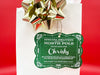 A green glitter cardstock Santa gift tag is shown hanging on a white gift bag. The bag is on a red backdrop and has a gold bow on it.