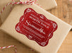 A red glitter cardstock Santa gift tag is shown on a craft paper wrapped box. The box also has a red and white striped string on it. The box is seen on a wooden surface.