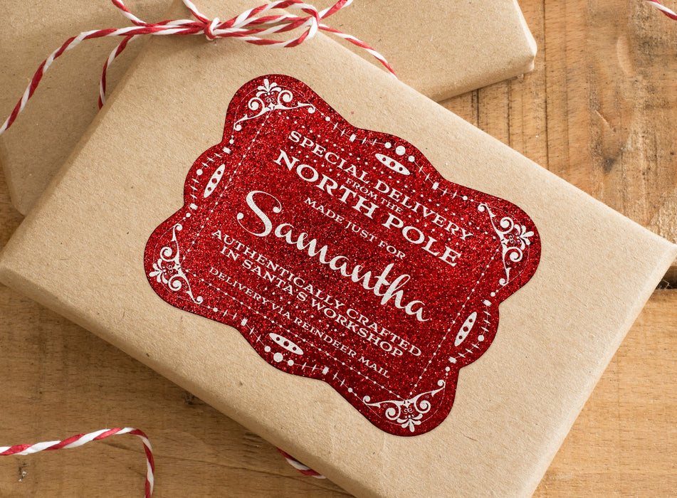 A red glitter cardstock Santa gift tag is shown on a craft paper wrapped box. The box also has a red and white striped string on it. The box is seen on a wooden surface.