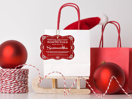 A red glitter cardstock Santa gift tag is shown hanging on a white gift bag. The bag is sitting on a sled and is surrounded by Christmas ornaments, a Santa hat, and a red gift bag. Everything is set against a white backdrop.