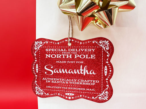 A red glitter cardstock Santa gift tag is shown hanging on a white gift bag. The bag is on a red backdrop and has a gold bow on it.