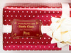 A gold foil cardstock Santa gift tag is shown on a Christmas present. The present paper is red with white polka dots. A white bow and ribbon are around the present. The box is seen on a white surface.