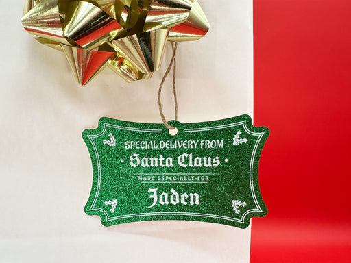 A green glitter cardstock Santa gift tag is shown hanging on a white gift bag. The bag is on a red backdrop and has a gold bow on it.