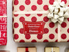 A red glitter cardstock Santa gift tag is shown on a Christmas present. The present paper is made of craft paper and has glittery red polka dots. A gold bow and ribbon are around the present. The box is seen on a white surface with other presents.