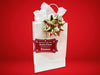 A red glitter cardstock Santa gift tag is shown hanging on a white gift bag. The bag is on a red backdrop and has a gold bow on it.