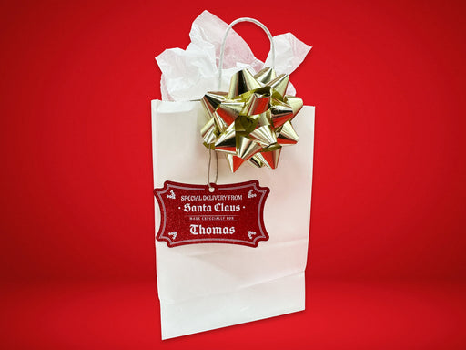 A red glitter cardstock Santa gift tag is shown hanging on a white gift bag. The bag is on a red backdrop and has a gold bow on it.